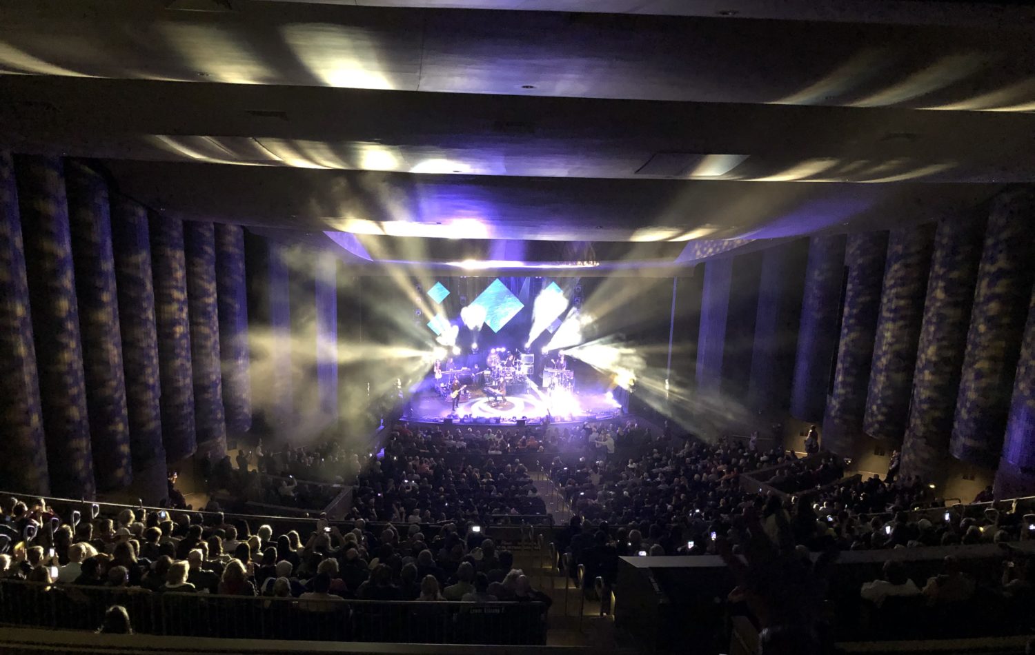 The Goo Goo Dolls perform on stage at the Mark C. Smith Concert hall. There is bright light coming from the stage and curtains on each side of the venue. There are people in the dark audience.