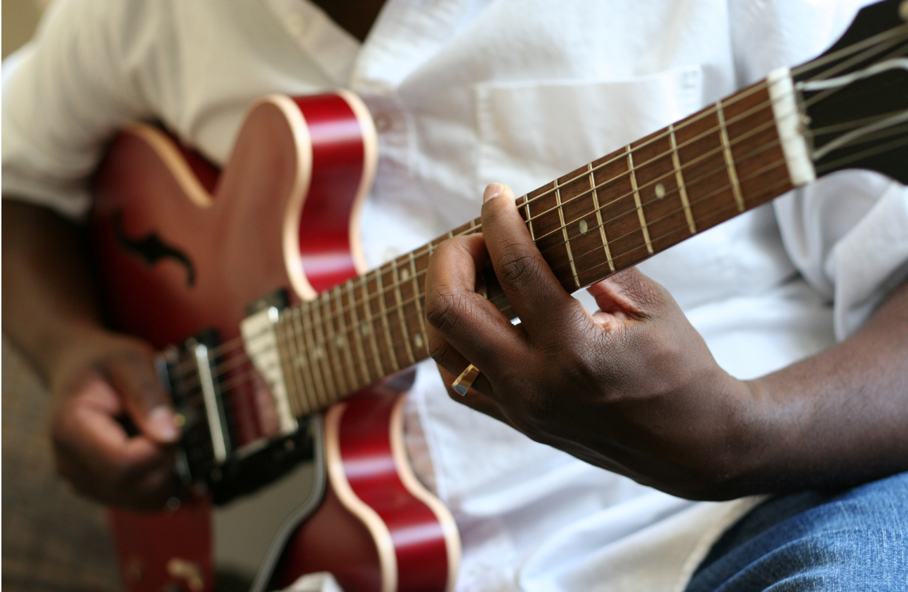 A man plays a red guitar. He's wearing a white shirt and blue jeans. His face is not visible.