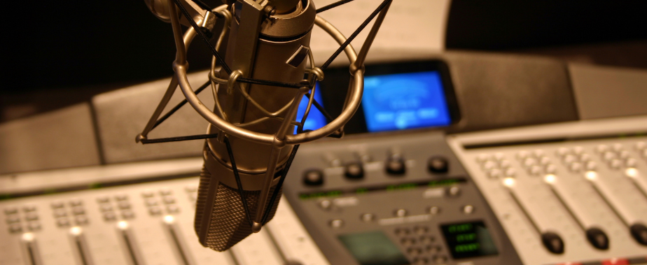 A microphone hangs in the foreground with a radio station console in the background. There are sliders and buttons and knobs and a blue screen.