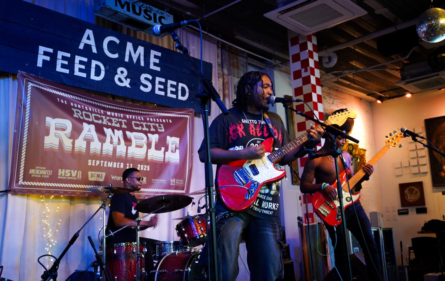 A three‑member band performs on stage at Acme Feed & Seed during the Rocket City Ramble event, with one guitarist at center playing a red electric guitar, another guitarist at right playing an orange guitar, and a drummer positioned behind them. A large event banner hangs in the background.